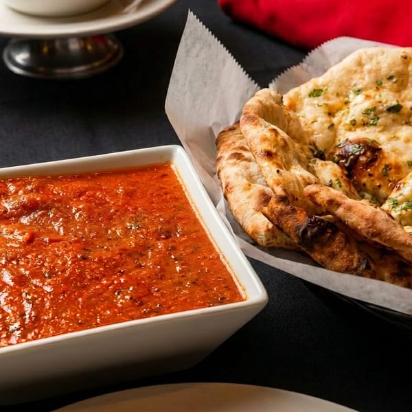 A white bowl of red curry sauce next to a basket of naan bread on a dark tablecloth.