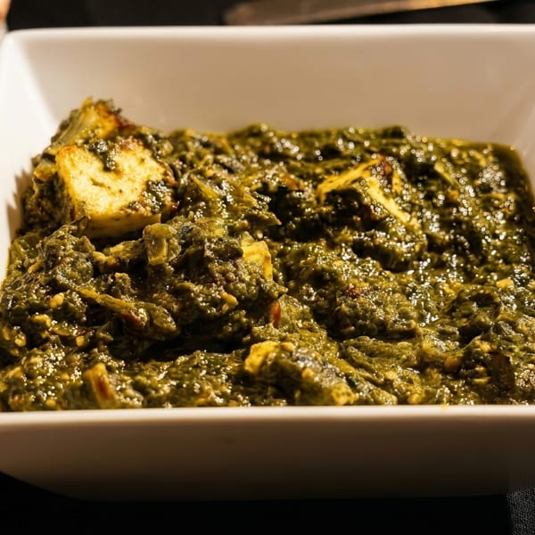 A white rectangular bowl filled with vibrant green palak paneer, with naan bread on the side.