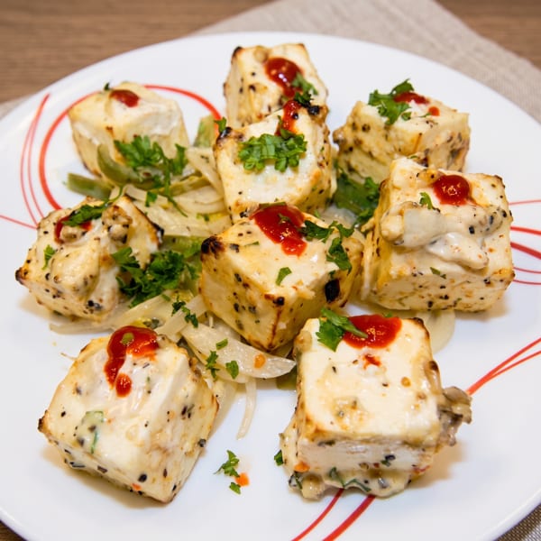 Eight pieces of paneer tikka, drizzled with red sauce and garnished with parsley, on a white plate.