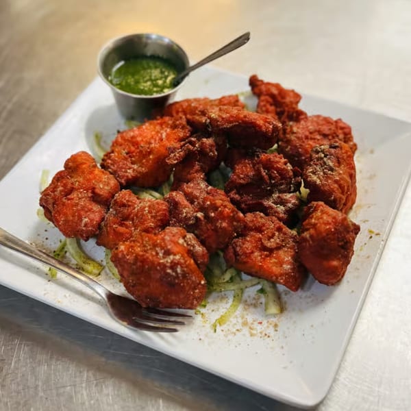 Crispy red chicken pakora served on a white plate with green chutney and a fork.