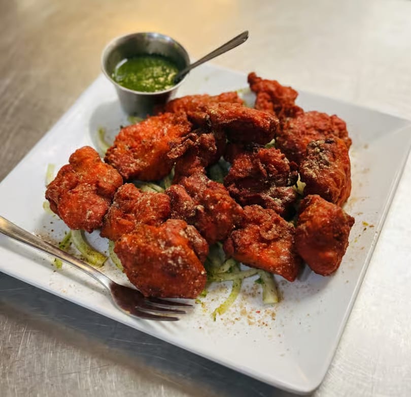 Crispy red chicken pakora served on a white plate with green chutney and a fork.