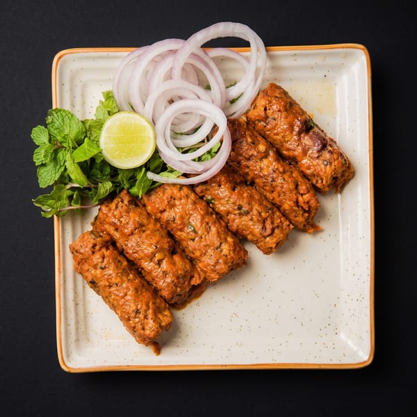 Six spicy seekh kebabs on a rectangular plate with mint, onion rings, and a lime wedge.